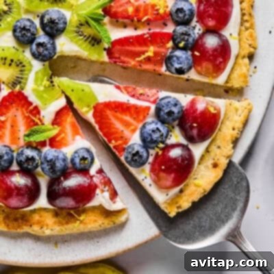 A slice of fruit pizza topped with blueberries, strawberries, grapes, kiwi, mint leaves, and lemon zest on a cookie crust with cream cheese frosting.