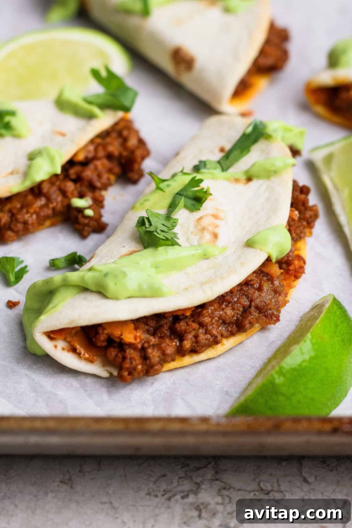 A perfectly cooked sheet pan taco filled with seasoned ground meat, topped with vibrant green avocado crema and fresh cilantro, served on a tray with bright lime wedges. The crispy tortilla texture is visible, inviting a delicious bite.