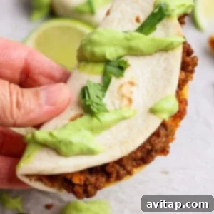 A hand holds a freshly baked and crispy sheet pan taco, garnished with creamy avocado crema and chopped cilantro, ready to be eaten.