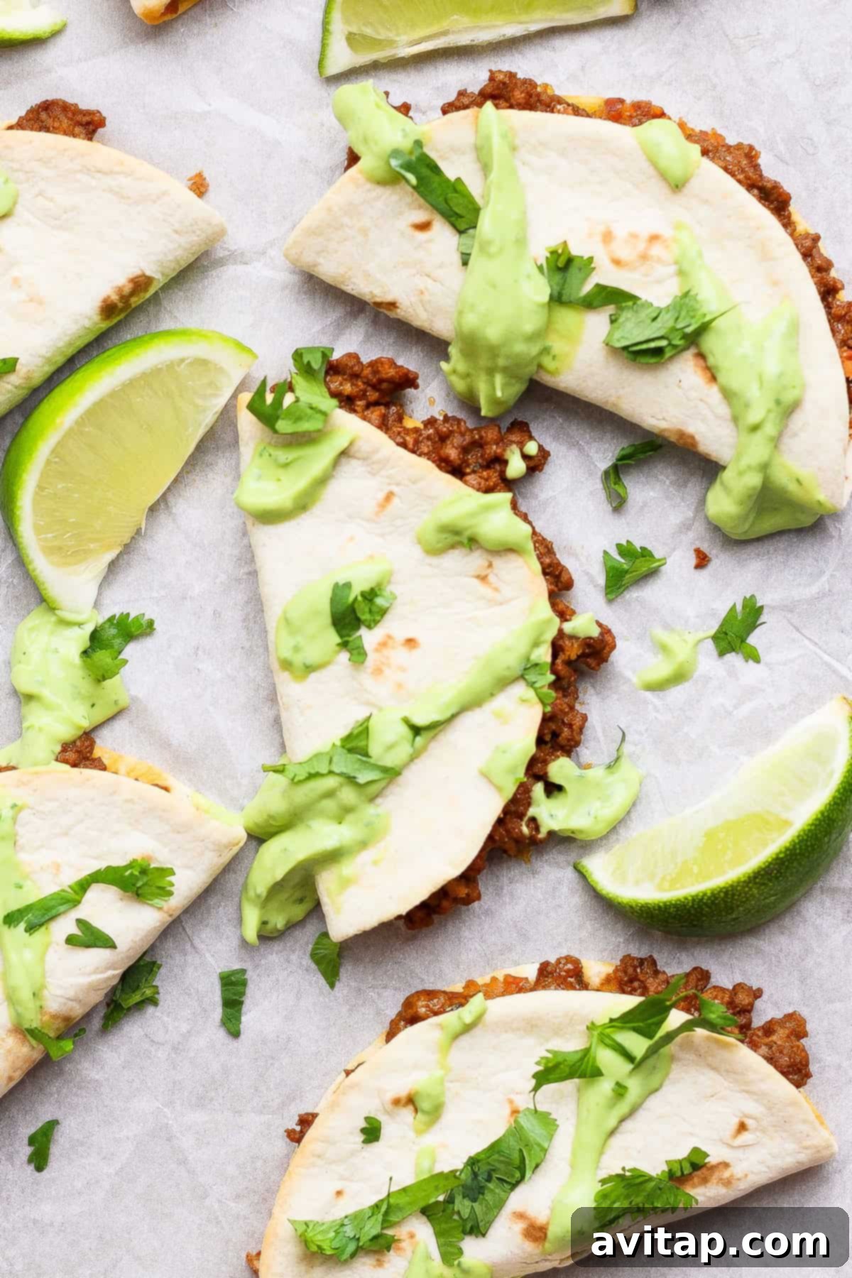 Three golden-brown folded sheet pan tacos, generously filled with seasoned ground beef, topped with creamy green avocado sauce and fresh cilantro, arranged on parchment paper with lime wedges on a rustic wooden board, ready for serving.