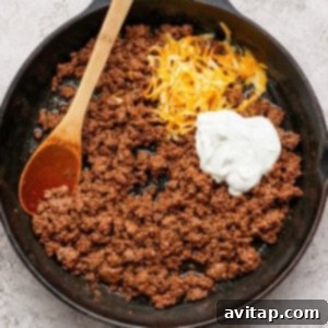 Ground beef cooking in a cast iron skillet with shredded Mexican blend cheese and a dollop of Greek yogurt being stirred in with a wooden spoon.