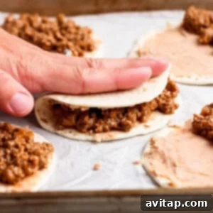 A hand gently folds a flour tortilla filled with ground beef and refried beans on a baking sheet lined with parchment paper, with other assembled tacos nearby.