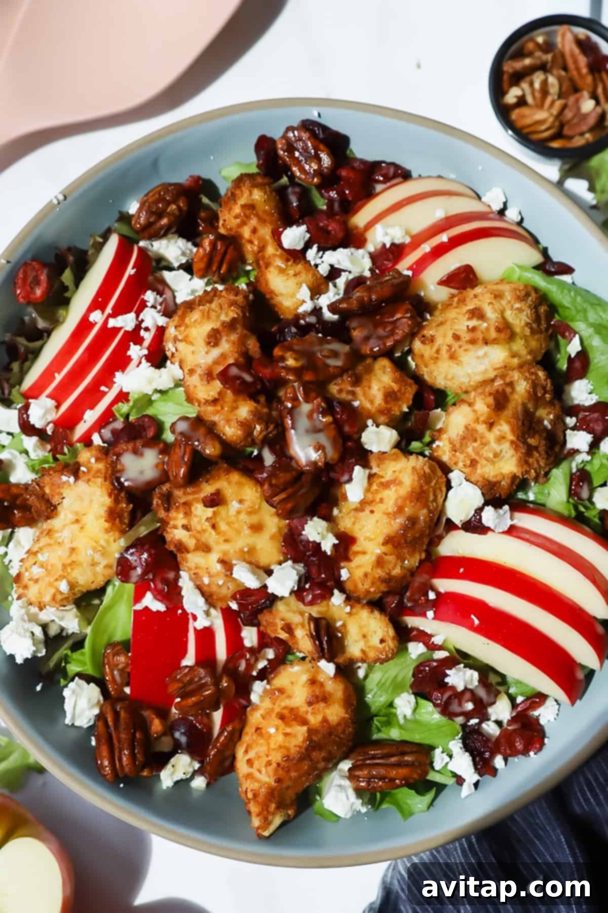 A vibrant, fresh salad in a gray bowl, featuring golden-brown breaded chicken pieces, thinly sliced red Honeycrisp apples, crunchy candied pecans, sweet dried cranberries, crisp mixed greens, and crumbled feta cheese, all beautifully drizzled with a creamy maple mustard dressing.