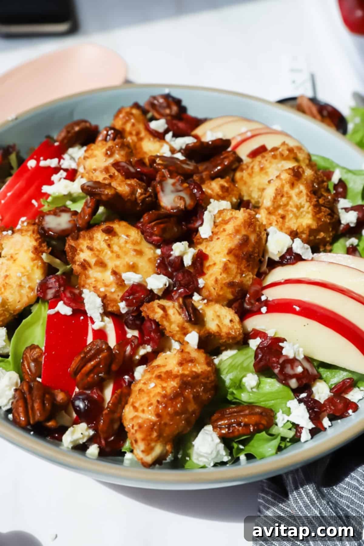 A close-up view of a hearty salad, showcasing mixed greens, golden-brown fried chicken pieces, thinly sliced red apples, crunchy pecans, tart dried cranberries, and crumbled feta cheese, with a light drizzle of dressing visible.