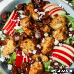 A vibrant salad with mixed greens, sliced red apples, crispy breaded chicken pieces, pecans, dried cranberries, crumbled feta cheese, and a light drizzle of dressing in a rustic bowl.