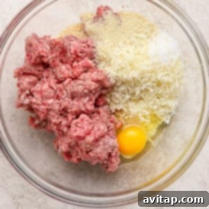 A glass bowl containing raw ground meat, grated cheese, breadcrumbs, and a whole egg on a light-colored surface.