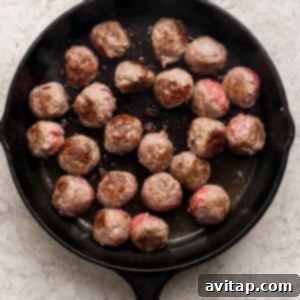 A black cast iron skillet with several beef meatballs being cooked, some browned and some still pink.