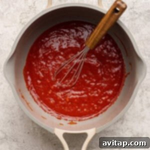 A saucepan filled with red tomato sauce and a metal whisk with a wooden handle resting inside, placed on a light-colored surface.