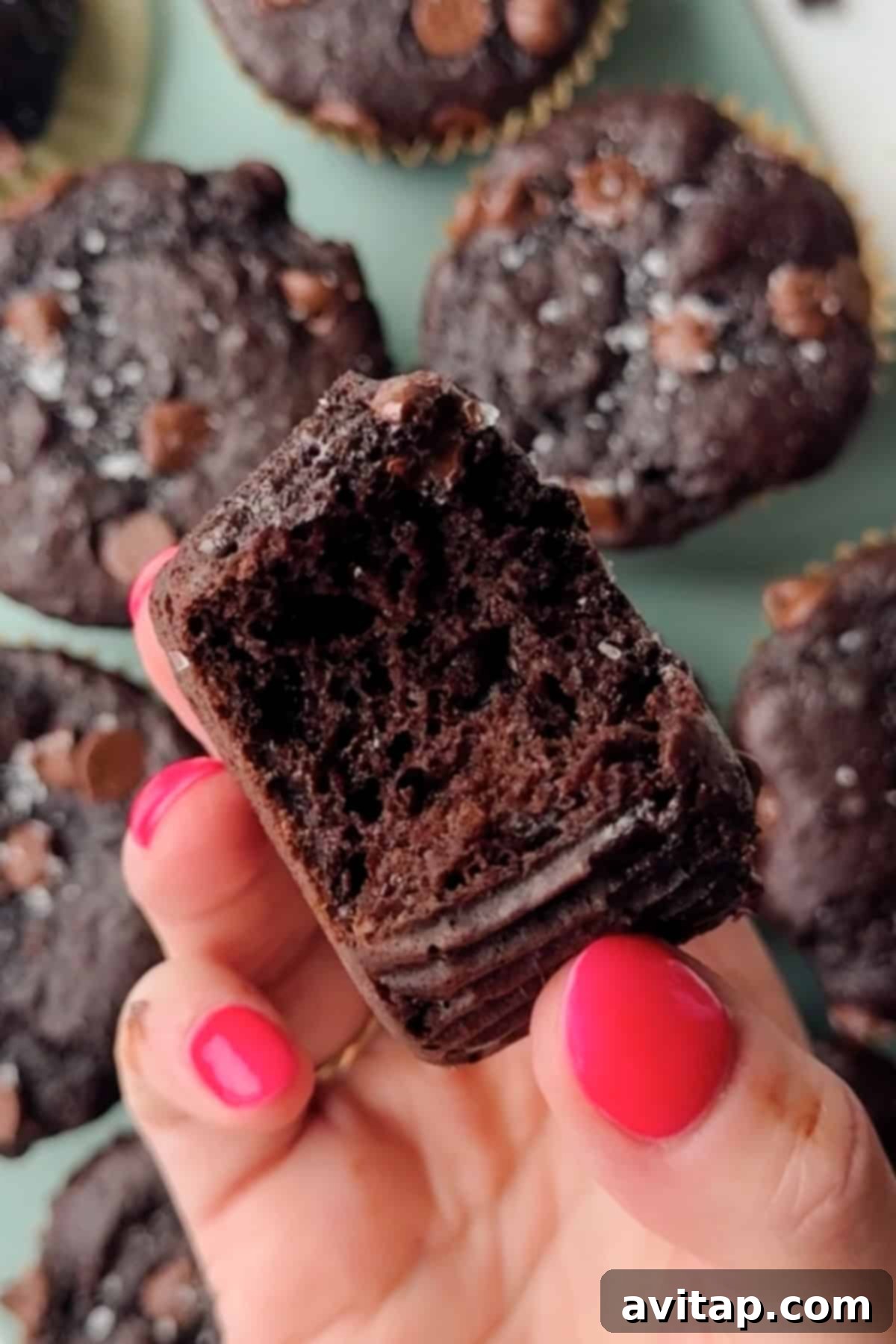 Protein-Packed Kodiak Chocolate Muffins 2 A hand with pink nail polish holds a partially eaten chocolate muffin, with more chocolate muffins visible in the background.