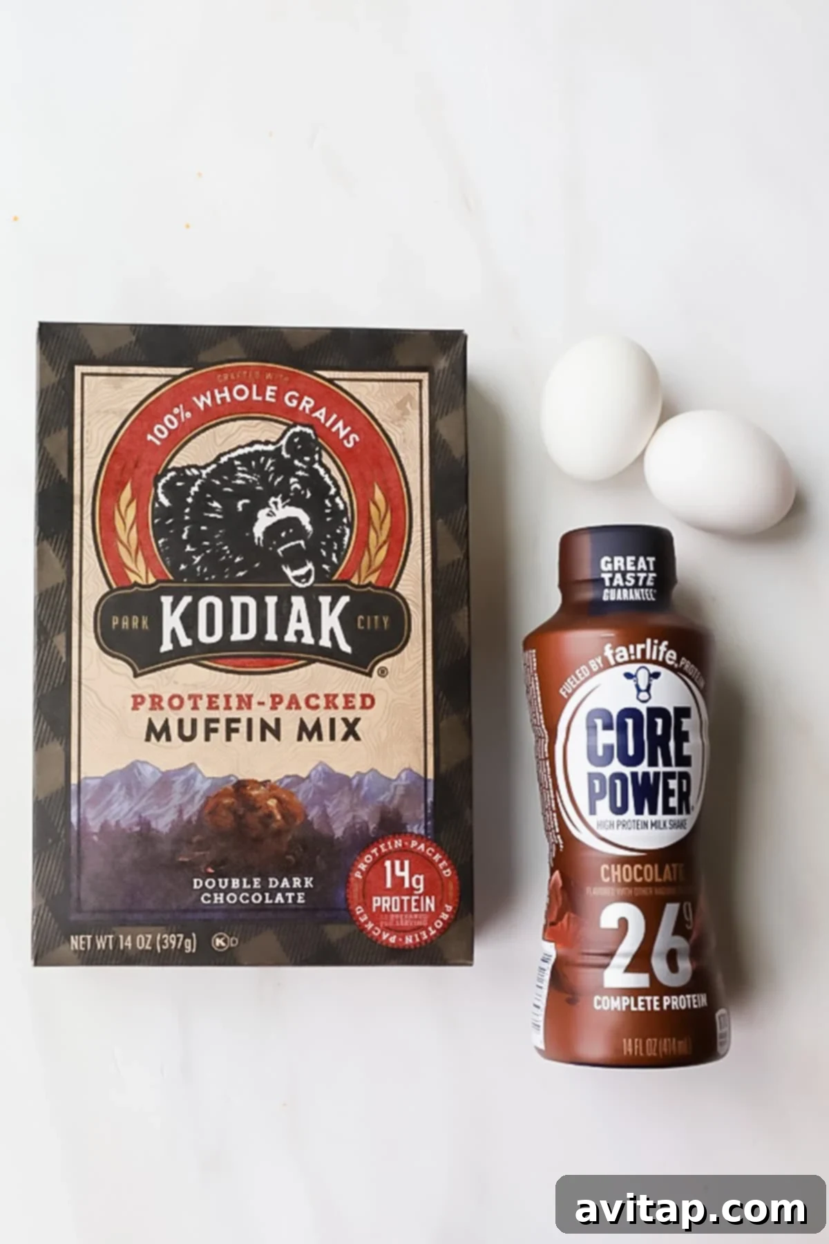 Protein-Packed Kodiak Chocolate Muffins 3 A box of Kodiak protein-packed muffin mix, a bottle of Core Power chocolate protein shake, and three white eggs are arranged on a white surface.