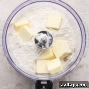 Butter chunks and flour in a food processor bowl, ready to be mixed, viewed from above on a light countertop.