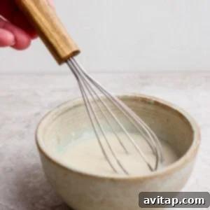 A hand holding a whisk stirring a light-colored mixture in a ceramic bowl on a light surface.