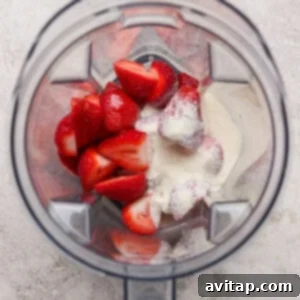 Overhead view of a blender containing halved strawberries and a scoop of white powder on a light textured surface.