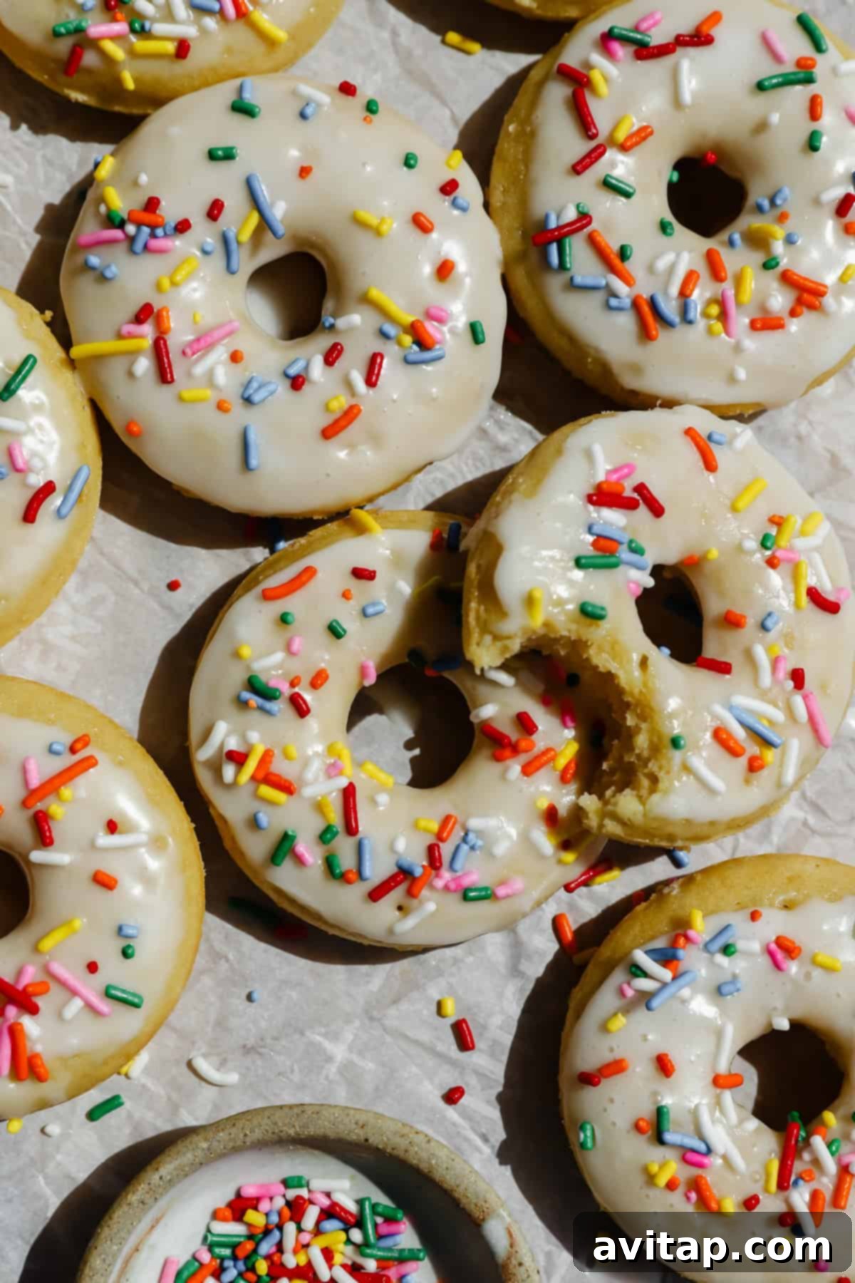 Beautifully glazed donuts topped with colorful sprinkles are artfully arranged on parchment paper, with one donut partially eaten, revealing its soft texture. A small bowl of extra sprinkles sits invitingly nearby.