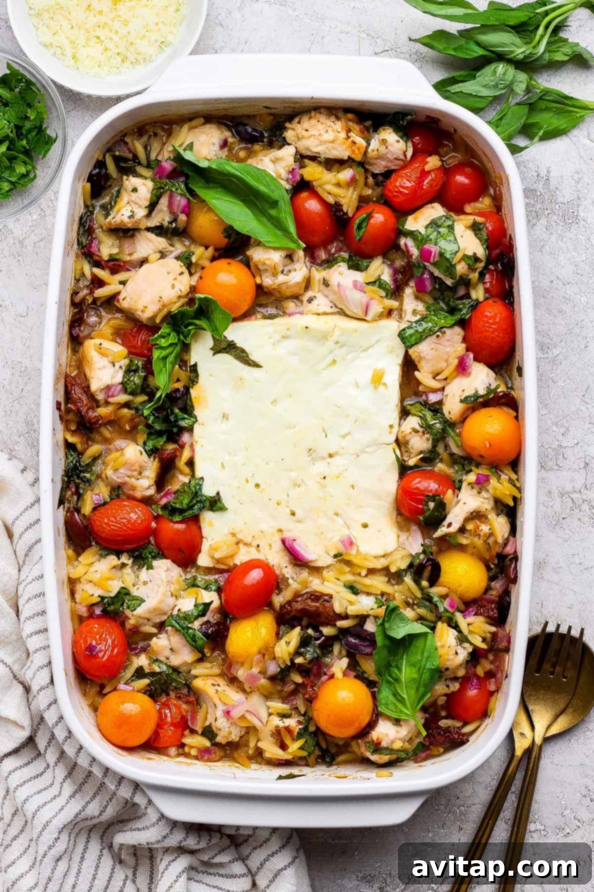 Savory Greek Chicken Orzo Bake 2 A baked casserole dish filled with chicken, cherry tomatoes, orzo pasta, red onions, and a large block of feta cheese in the center, garnished with fresh basil.