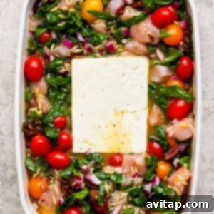 Savory Greek Chicken Orzo Bake 10 A white baking dish filled with raw chicken pieces, cherry tomatoes, spinach, chopped red onions, orzo, and a large block of feta cheese in the center.