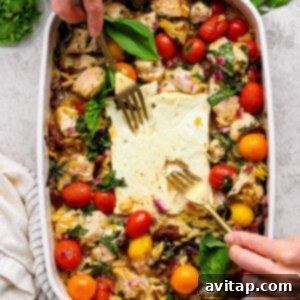Savory Greek Chicken Orzo Bake 11 A person uses two forks to serve a baked dish with chicken, cherry tomatoes, greens, orzo, and feta cheese in a white casserole dish. Fresh herbs are visible on the side.