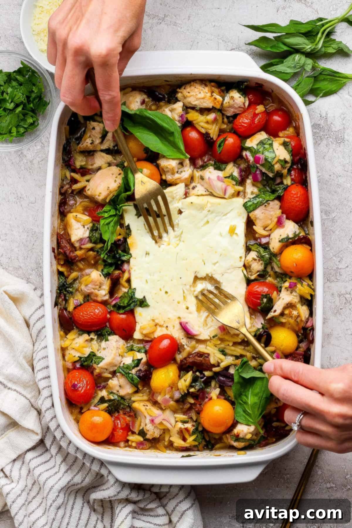 Savory Greek Chicken Orzo Bake 5 A person uses two forks to serve a baked dish with chicken, cherry tomatoes, greens, orzo, and feta cheese in a white casserole dish. Fresh herbs are visible on the side.