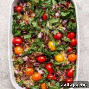 Savory Greek Chicken Orzo Bake 7 A white baking dish filled with fresh spinach, cherry tomatoes, chopped red onion, sun-dried tomatoes, and olives on a light textured surface.