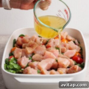 Savory Greek Chicken Orzo Bake 9 A hand pours broth from a glass measuring cup over raw chopped chicken, tomatoes, and greens in a white baking dish.