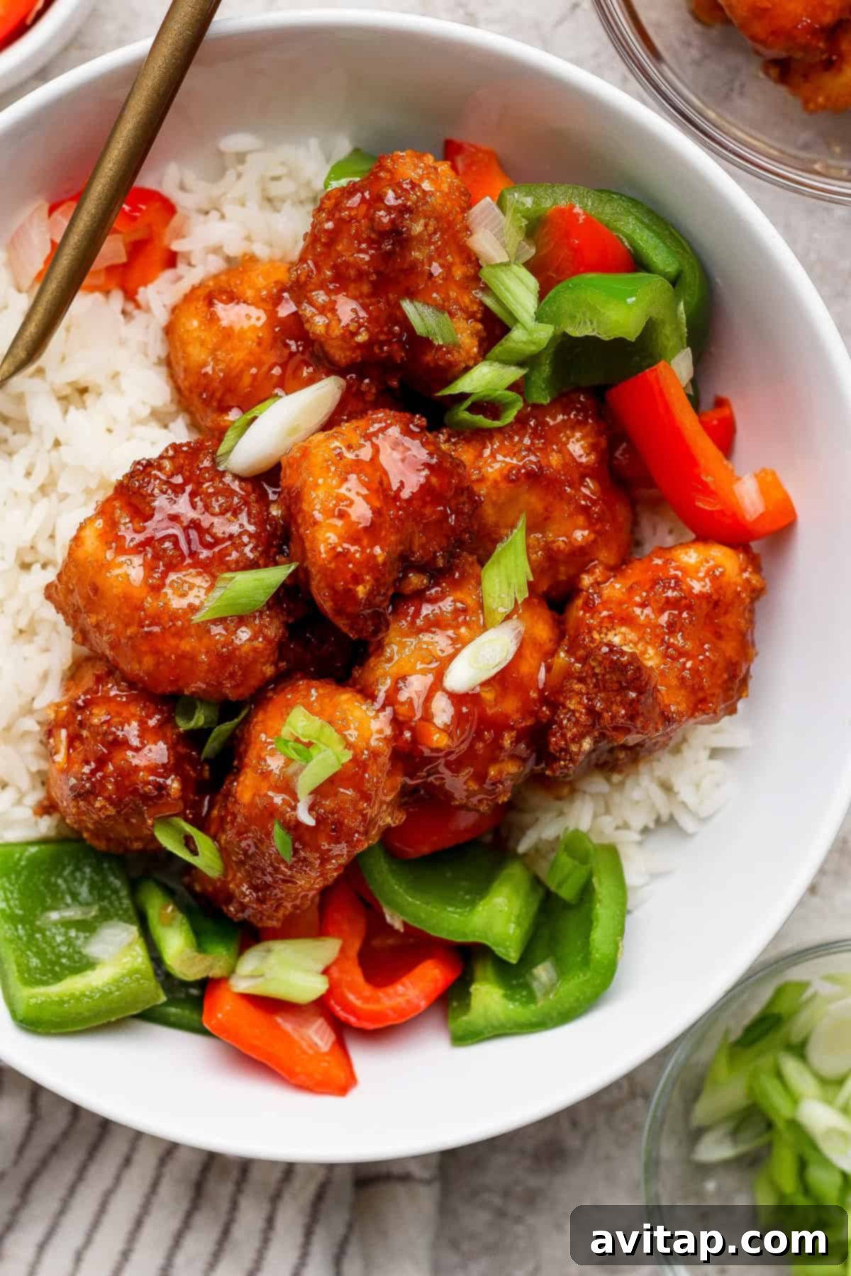 A beautifully presented bowl of white rice topped with generous portions of glazed, crispy air fryer sweet and sour chicken, adorned with freshly chopped green onions, and colorful sliced red and green bell peppers.