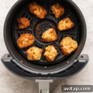 Eight breaded chicken nuggets are arranged in the basket of an air fryer, cooked to a golden brown color on a textured light surface.