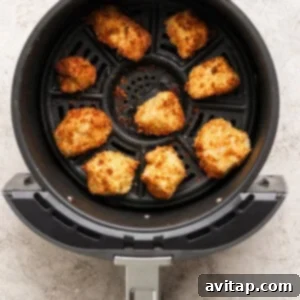 Eight breaded chicken nuggets are arranged in the basket of an air fryer, cooked to a golden brown color on a textured light surface.