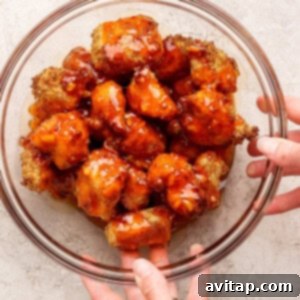 A clear glass bowl filled with breaded, sauce-coated cauliflower bites is being held by two hands over a light-colored surface.