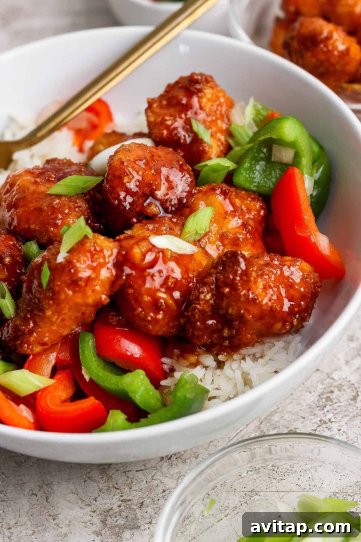 A beautifully presented bowl of white rice topped with generous portions of crispy glazed sweet and sour chicken, vibrant sliced red and green bell peppers, and a sprinkle of fresh chopped green onions.