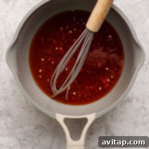A saucepan containing a reddish-brown sauce with chili flakes, being whisked with a metal whisk on a textured light surface.