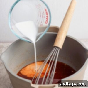 A hand pours a white liquid from a measuring cup into a pot containing a brown sauce, with a whisk resting inside the pot.