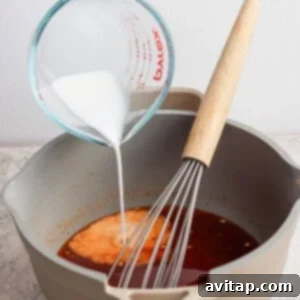 A hand pours a white liquid from a measuring cup into a pot containing a brown sauce, with a whisk resting inside the pot.