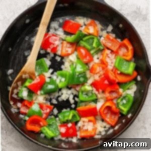 A cast-iron skillet with chopped red and green bell peppers and diced onions being sautéed, stirred with a wooden spoon.