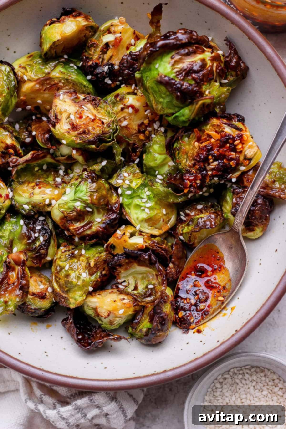 Addictive Chili Crunch Brussels Sprouts 2 A vibrant bowl of perfectly roasted Brussels sprouts, generously garnished with white sesame seeds and drizzled with a rich chili oil, served with a spoon on a rustic surface. This delectable side dish showcases its crispy texture and enticing aroma.