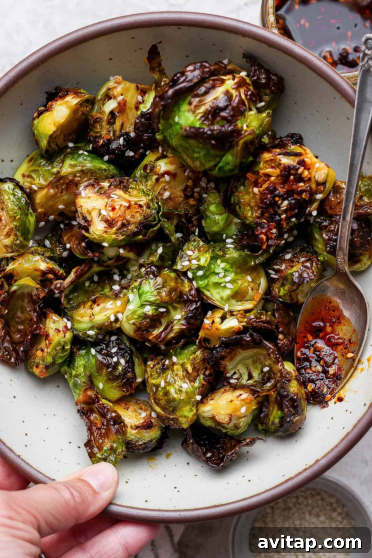 Addictive Chili Crunch Brussels Sprouts 4 A close-up shot of a bowl filled with delectable roasted Brussels sprouts, liberally sprinkled with white sesame seeds and a glossy hint of chili oil. A serving spoon rests against the side, ready to scoop these irresistible, crispy bites.