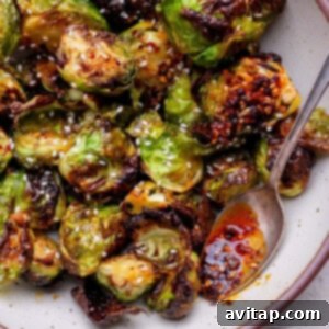Roasted Brussels sprouts with a glossy chili sauce, garnished with sesame seeds, served in a bowl with a spoon.