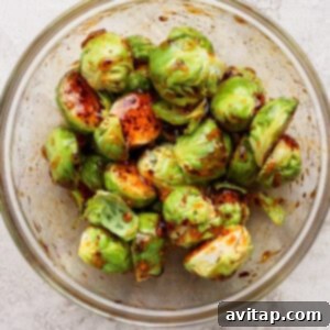 Addictive Chili Crunch Brussels Sprouts 7 A glass bowl filled with halved Brussels sprouts coated in a dark seasoning or marinade, placed on a light textured surface.