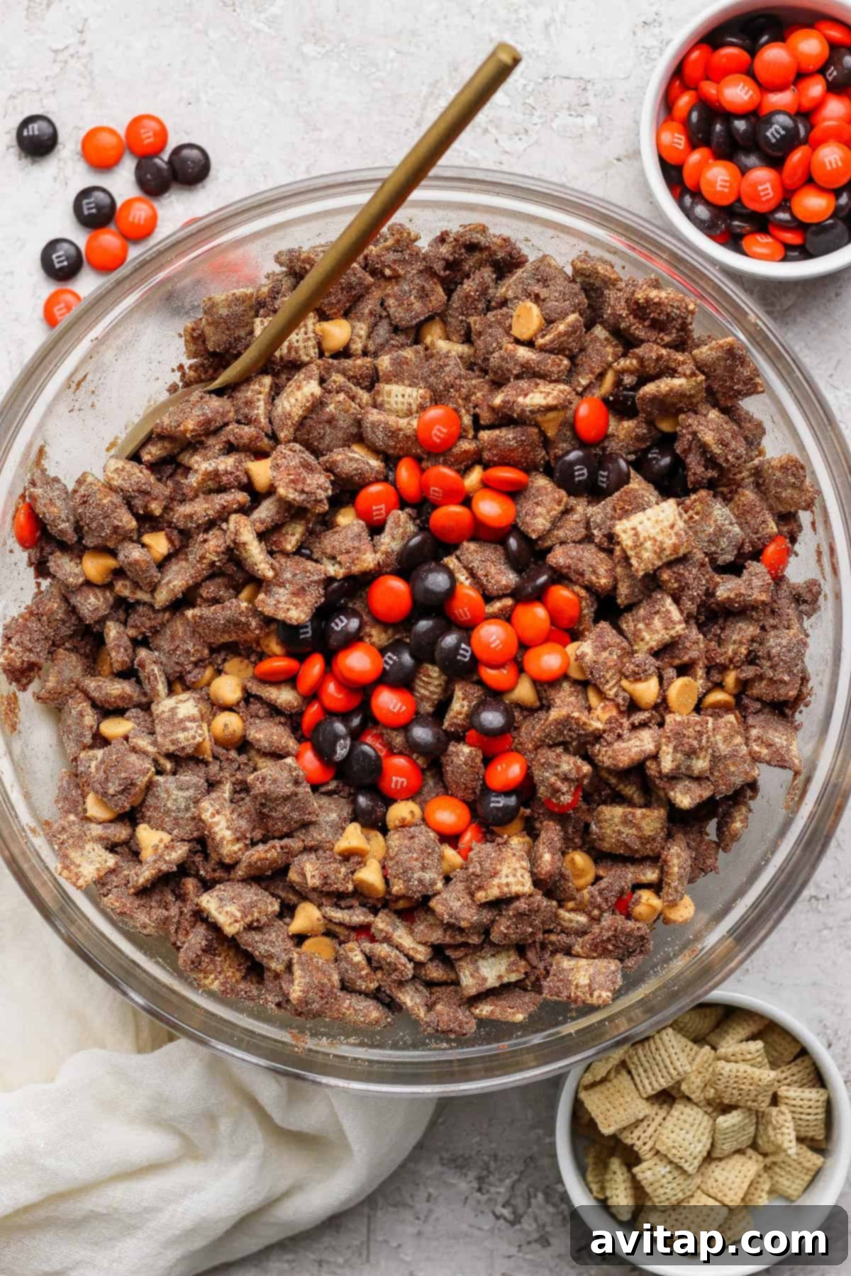 A beautiful glass bowl overflowing with chocolate-coated Chex cereal, interspersed with rich peanut butter chips, and festive orange and brown M&M candies. Small side bowls holding extra M&Ms and plain Chex cereal are artfully arranged nearby.