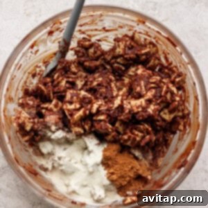 A clear glass bowl contains chocolate-coated Chex cereal, generously dusted with white powdered sugar (representing protein powder) and brown cinnamon (pumpkin spice), with a gray spatula poised inside for mixing.