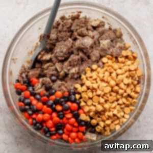 A clear glass bowl filled with chocolate-covered cereal, vibrant peanut butter chips, and a festive mix of red and brown candy-coated chocolates, with a gray spoon resting inside, all ready for the final stir.