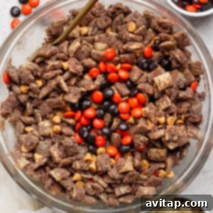 A spacious glass bowl brimming with chocolate-coated Chex cereal, adorned with vibrant peanut butter chips, and festive orange and brown M&M candies. Smaller bowls containing extra M&Ms and plain Chex are placed around it, showcasing the finished product ready to serve.