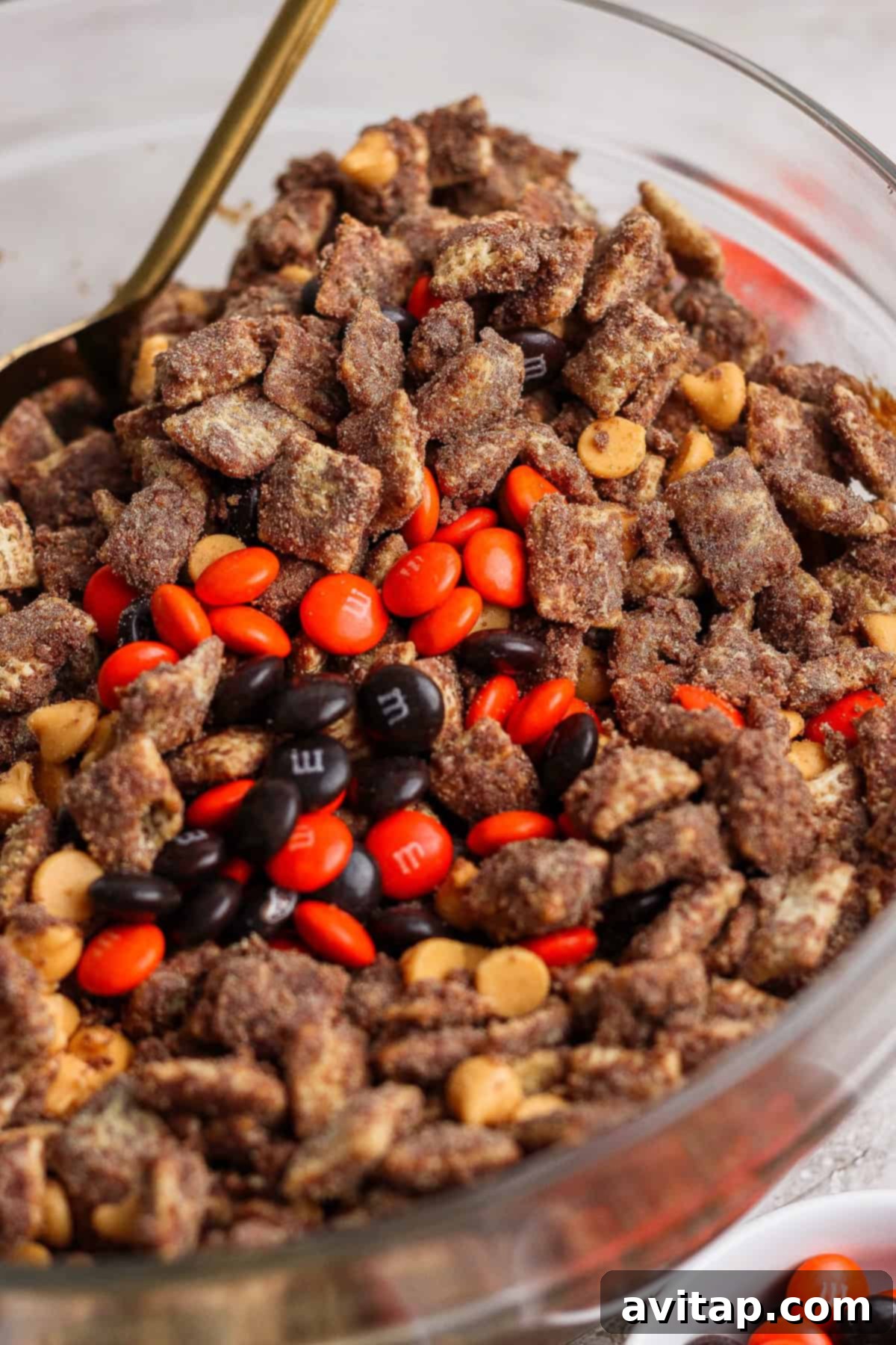 A crystal-clear glass bowl brimming with chocolate-coated cereal pieces, artfully mixed with vibrant peanut butter chips and an assortment of red, orange, and brown candy-coated chocolates, showcasing the finished protein puppy chow.