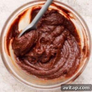 A clear glass bowl contains a rich, thick chocolate mixture ready to be incorporated into the puppy chow, with a gray spatula resting inside, all on a light textured surface.