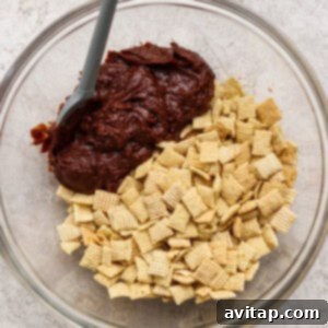 A glass bowl holds a mix of crunchy Chex cereal and melted chocolate, combined with a spatula on a textured light surface, illustrating the initial coating process.