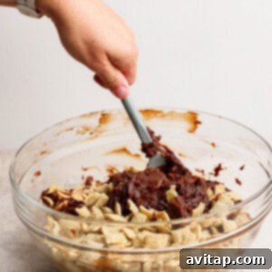 A person is actively mixing chocolate with Chex cereal in a transparent glass bowl using a spatula, capturing the detailed process of creating the snack.