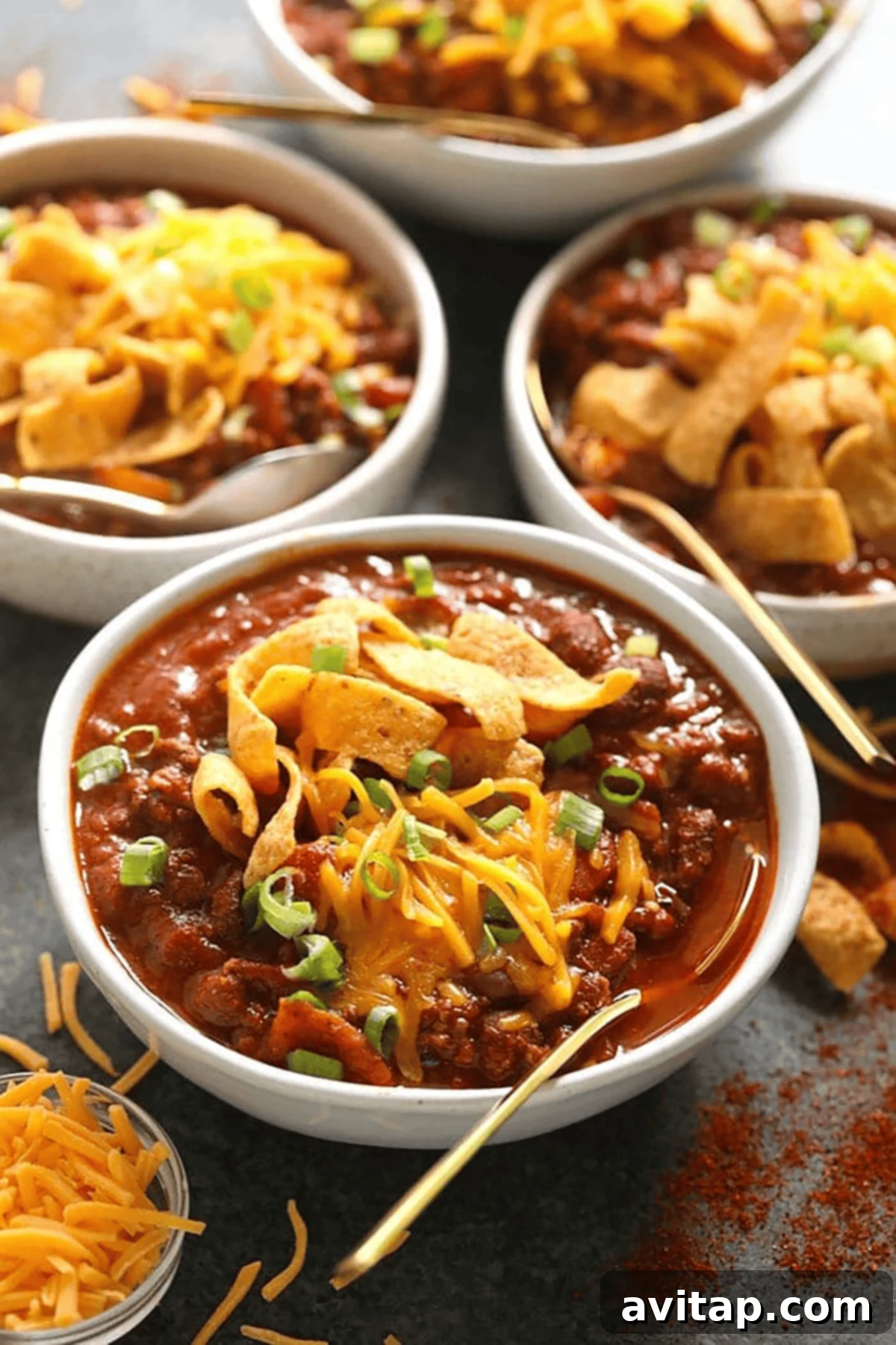 Championship Chili Recipe 2 Four bowls of chili topped with shredded cheddar cheese, corn chips, and chopped green onions, with a side of shredded cheese and gold spoons. A perfect presentation of a winning chili recipe.