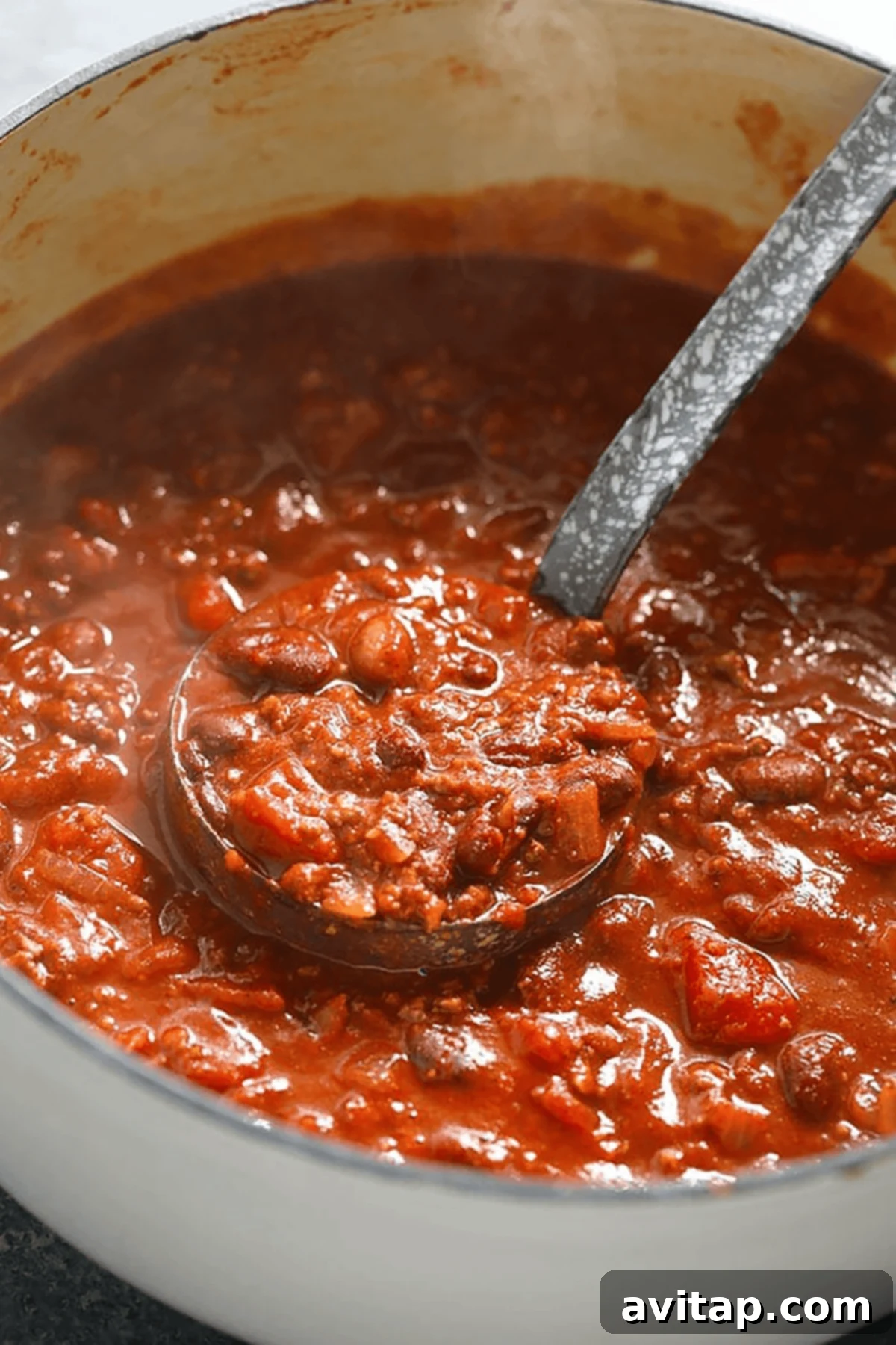Championship Chili Recipe 3 A ladle scoops thick chili with beans, tomato chunks, and ground meat from a large white pot—an inviting chili recipe in progress, showcasing its rich texture.