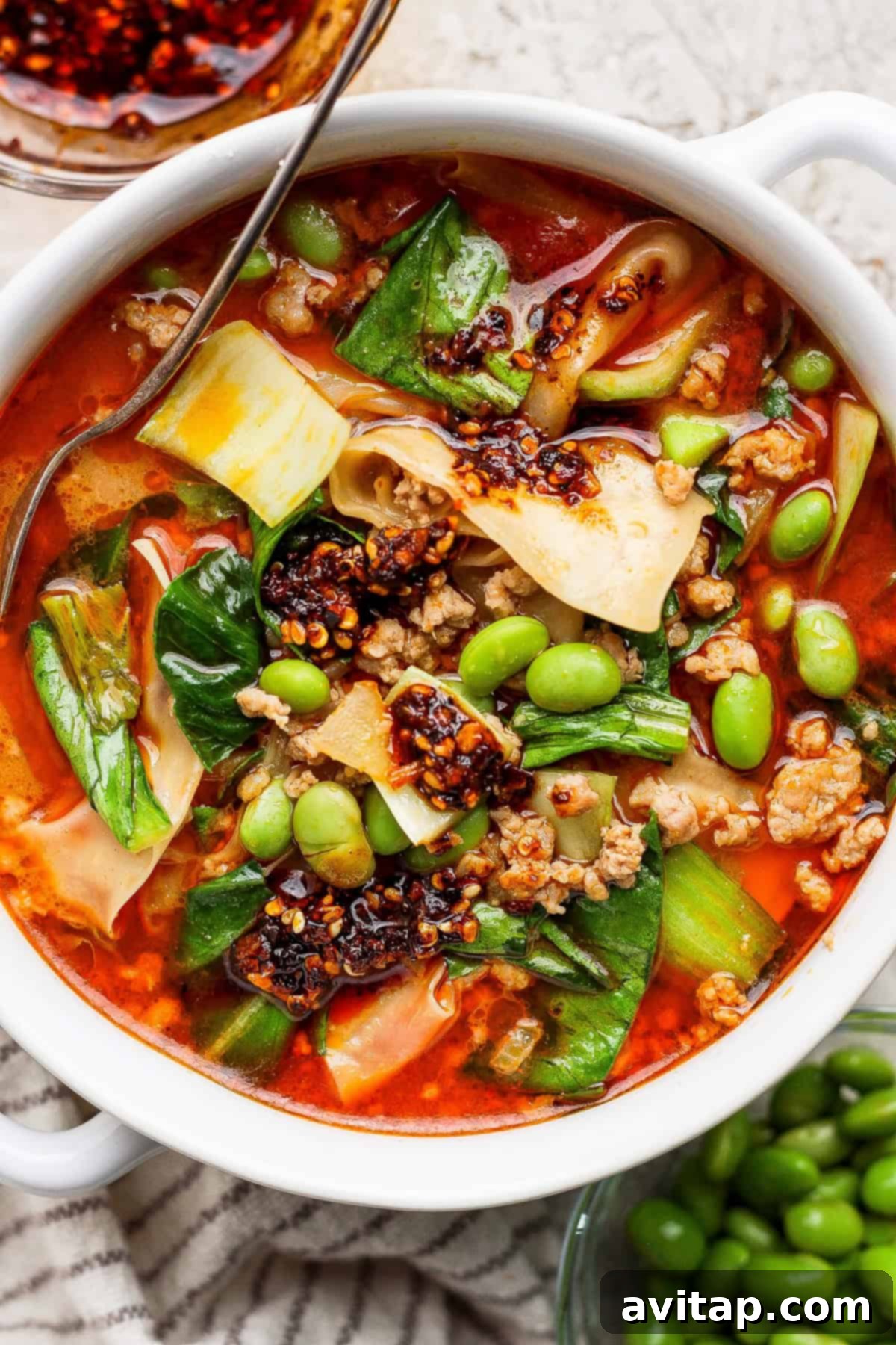 A vibrant bowl of Thai Coconut Dumpling Soup featuring deconstructed pork dumplings, wide noodles, green bok choy, and edamame, garnished with chili oil, served with a metal spoon on a rustic wooden surface.