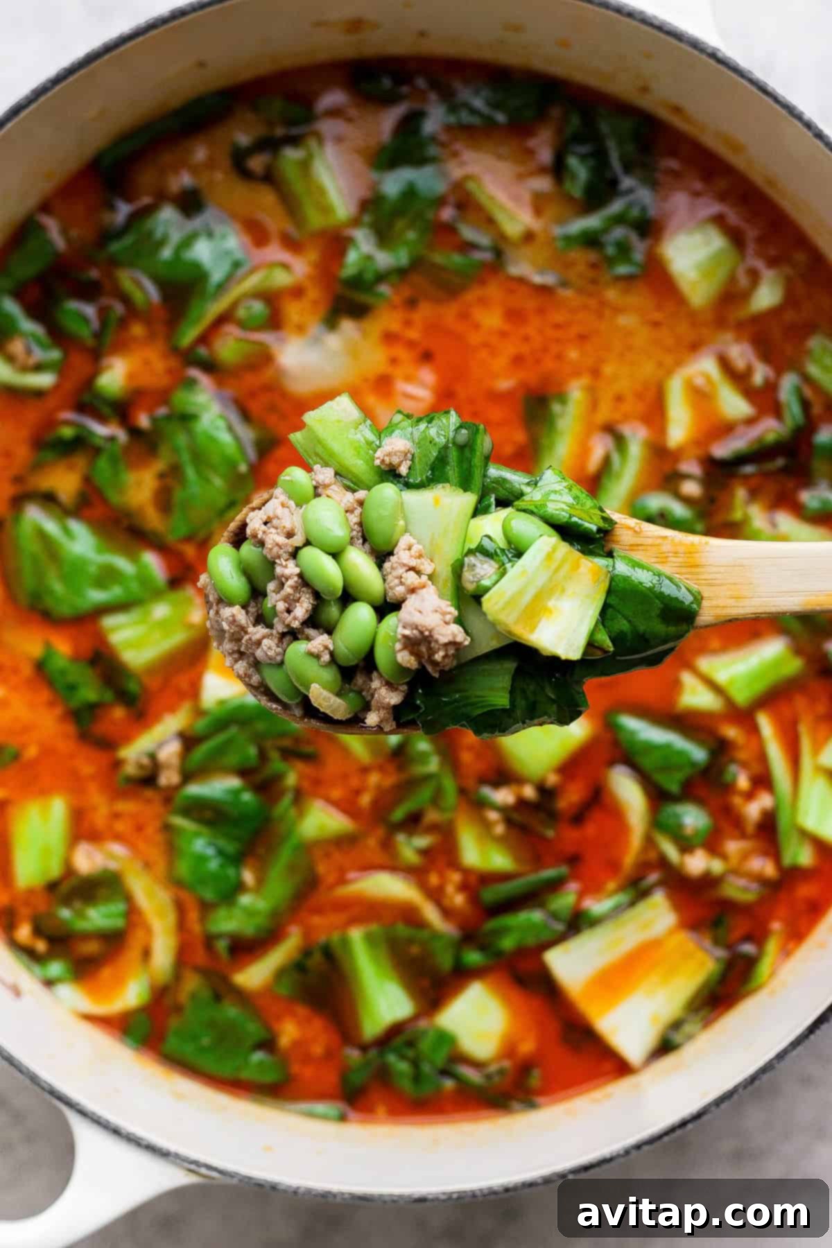 A wooden spoon lifting a spoonful of Thai Coconut Dumpling Soup from a pot, showing ground meat, edamame, and bok choy in a rich, red broth.
