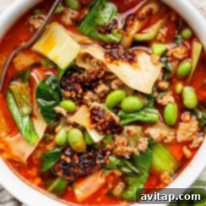 A bowl of spicy noodle soup with ground meat, bok choy, edamame, chili oil, and wide noodles, with a spoon partially visible.